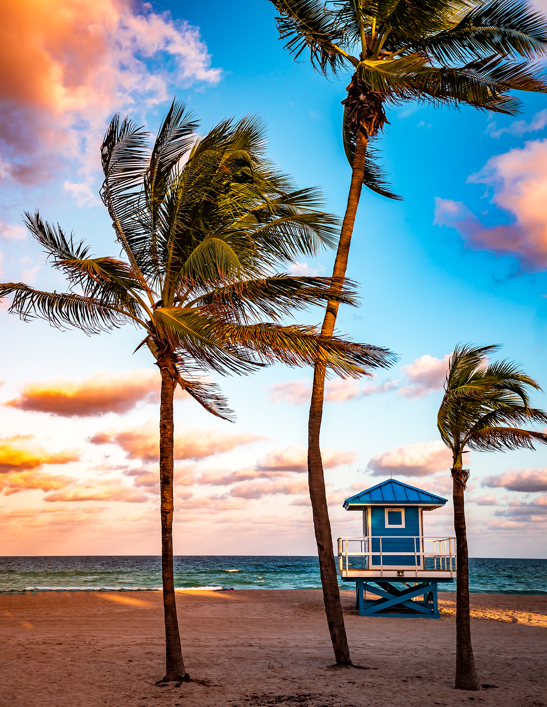 Sunset over a sandy beach with palm trees and a blue lifeguard tower by the sea.